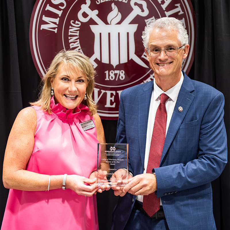 Two people smiling and holding an award in front of a university logo backdrop. The person on the left is wearing a pink dress, and the person on the right is in a blue suit and red tie.