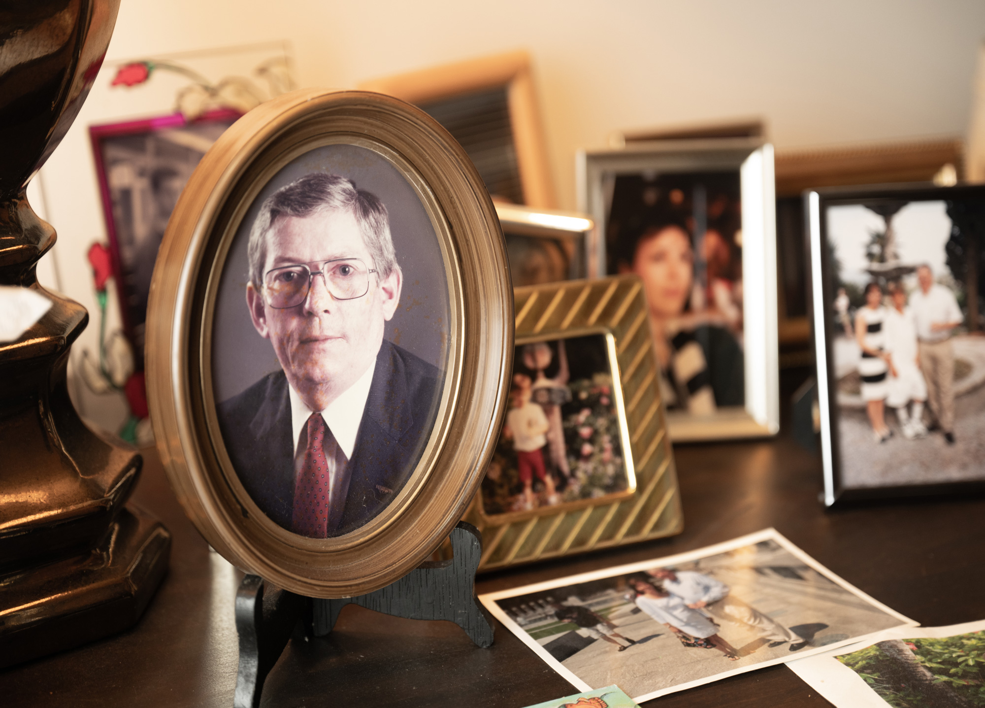 A collection of framed photographs on a table, featuring portraits of people in various settings, with a prominent circular framed portrait of an older man wearing glasses and a suit.