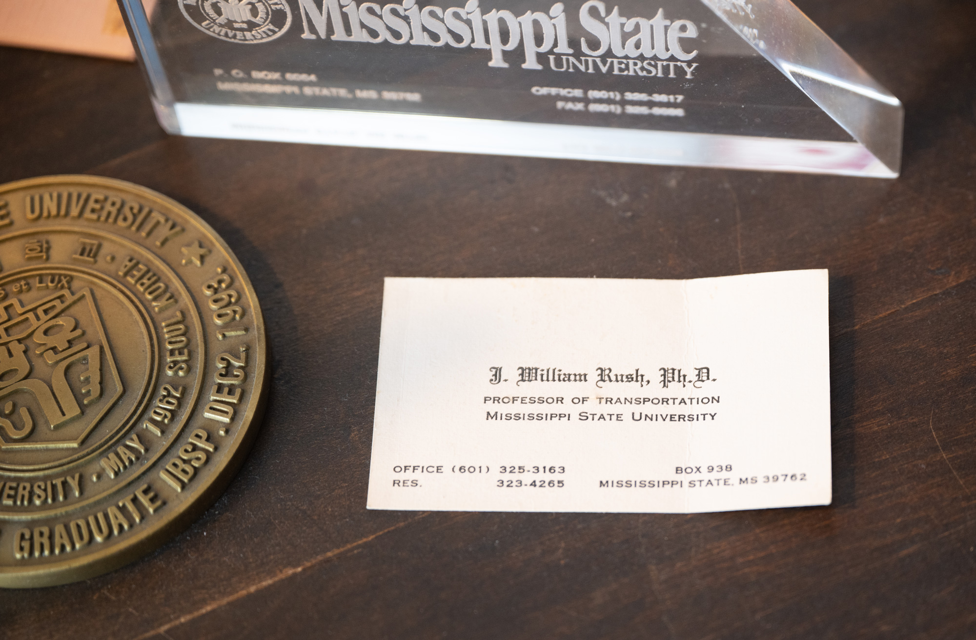 A business card for J. William Rush, Ph.D., Professor of Transportation at Mississippi State University, lies on a dark surface, with a university plaque and clear award nearby.