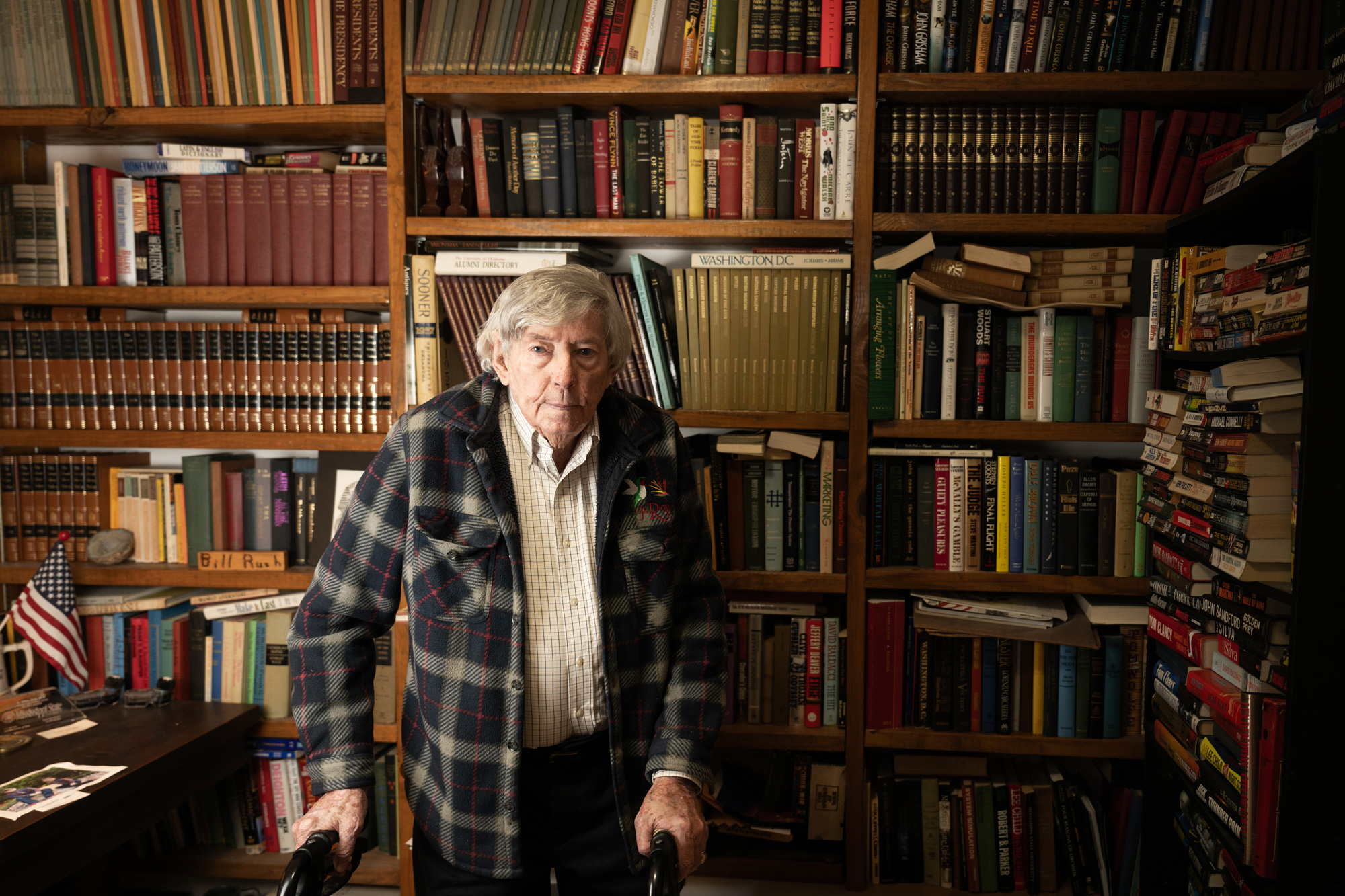 An elderly man stands with a walker in a room lined with bookshelves filled with various books. A desk with papers and small American flag is visible on the left.