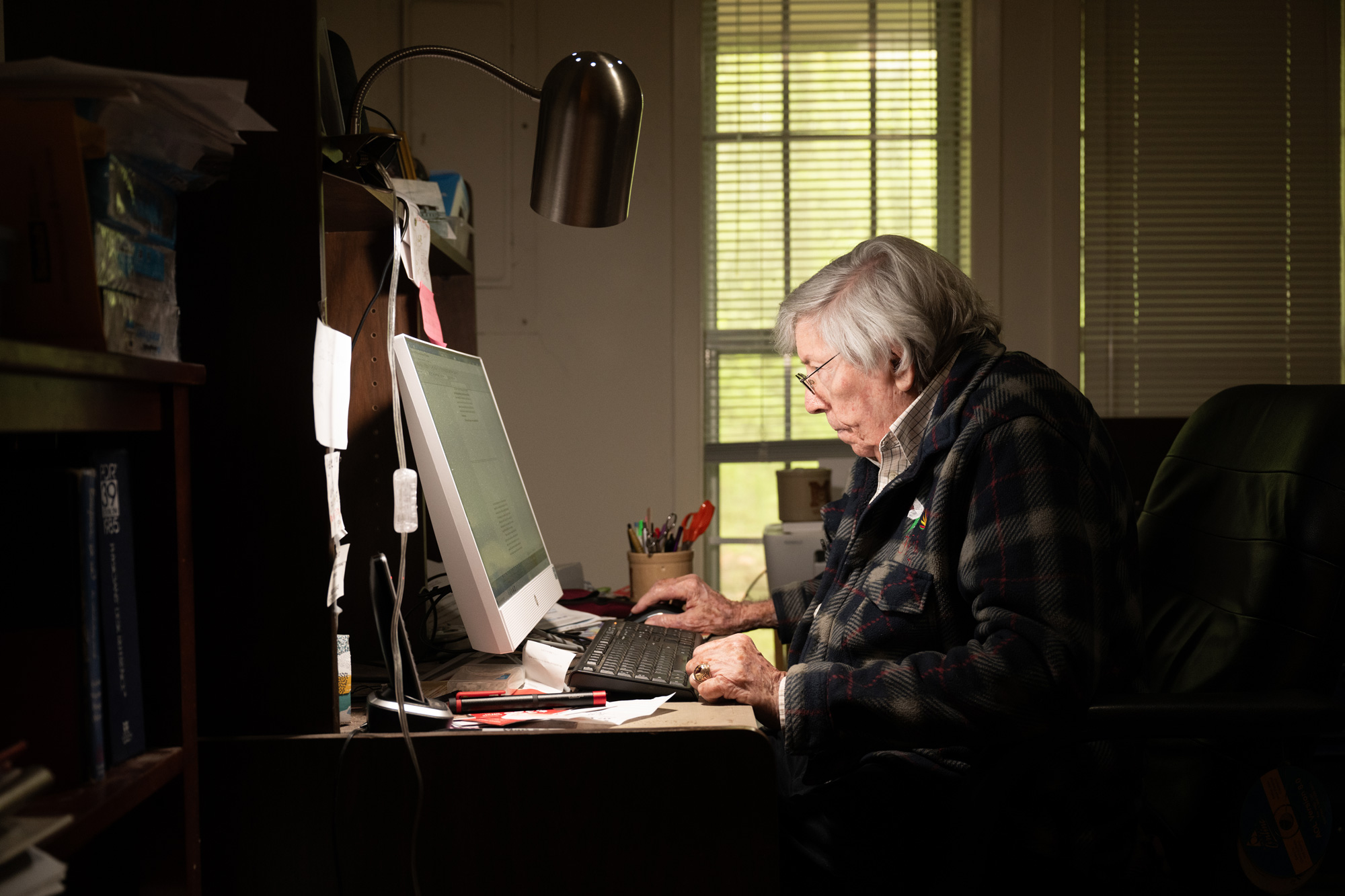 An elderly person with gray hair is focused on using a computer at a desk in a dimly lit room.