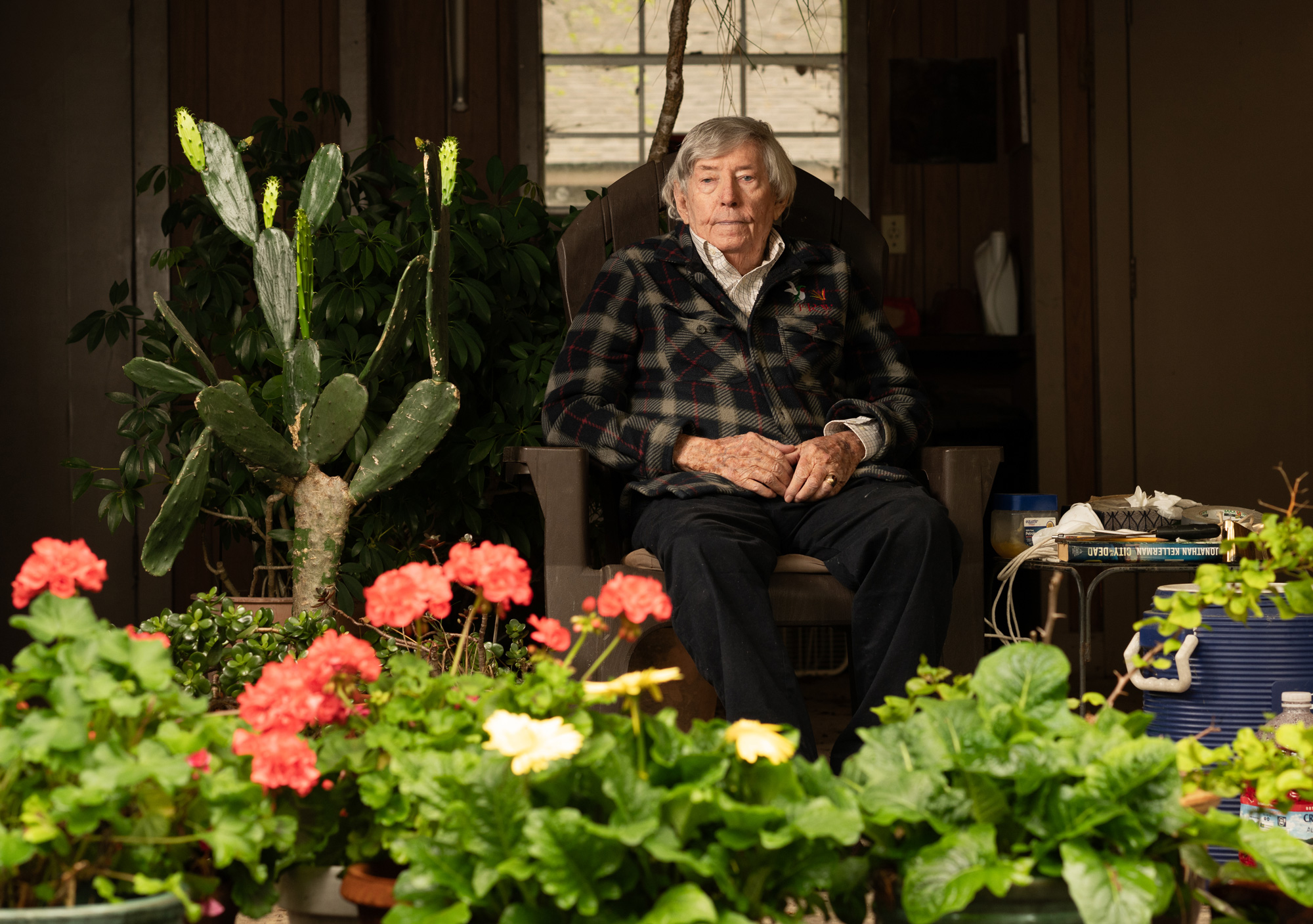 An older man sits on a chair in a room surrounded by various plants, including cacti and blooming flowers.