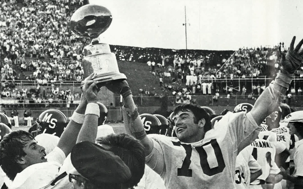 Football players celebrate with a trophy on the field, surrounded by a cheering crowd.