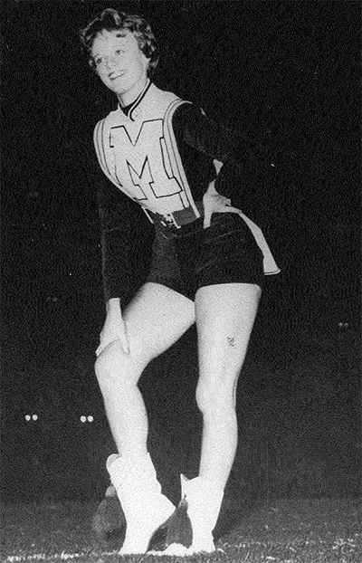 Black and white photo of a cheerleader posing on a field at night, wearing a uniform with the letter "M.