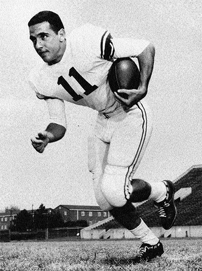 A football player in a white uniform with number 11 runs on a field holding a football. The background shows buildings and bleachers.