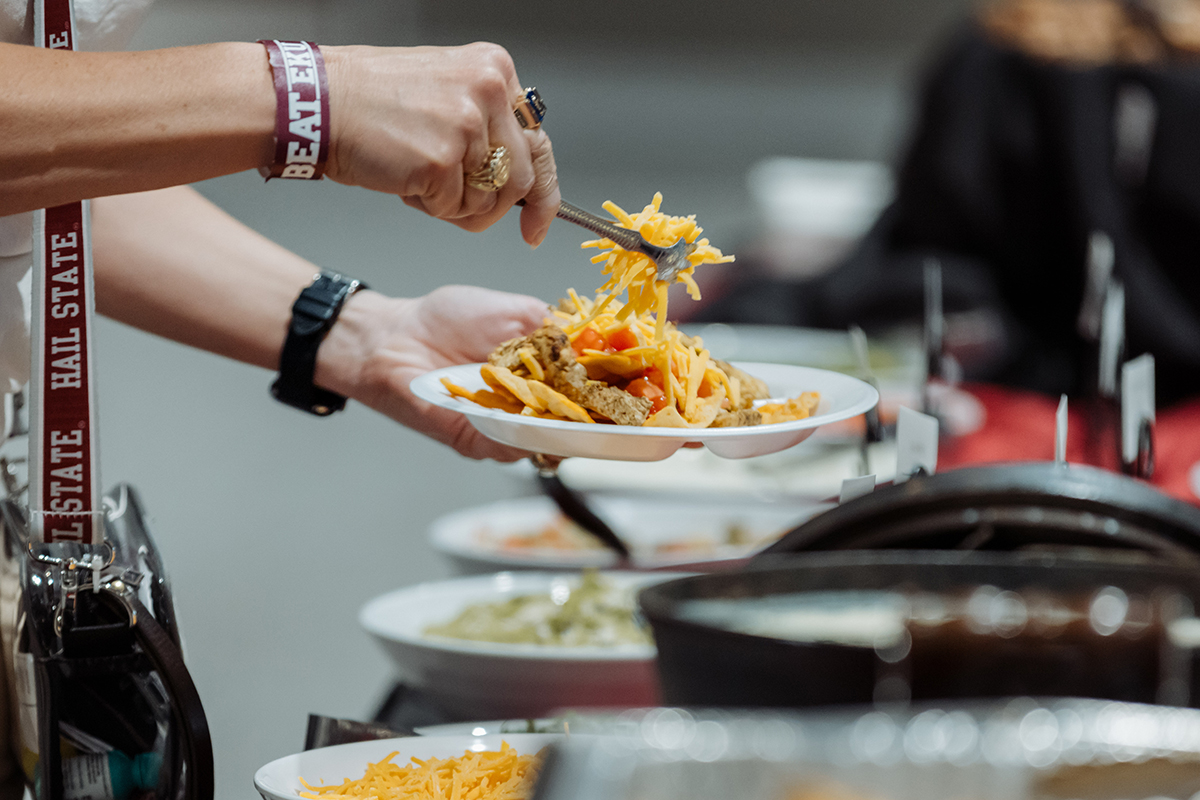 Person serving themselves a plate of pasta from a buffet, with various toppings and dishes visible in the background.