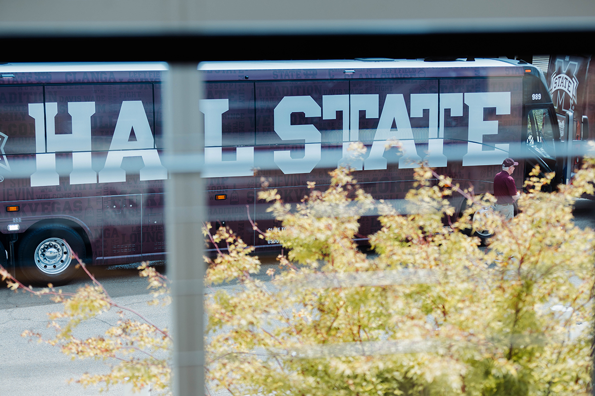 Bus with "HAIL STATE" written on the side is parked, with a person in maroon clothing standing next to it. Bushes with green and yellow leaves partially visible in the foreground.