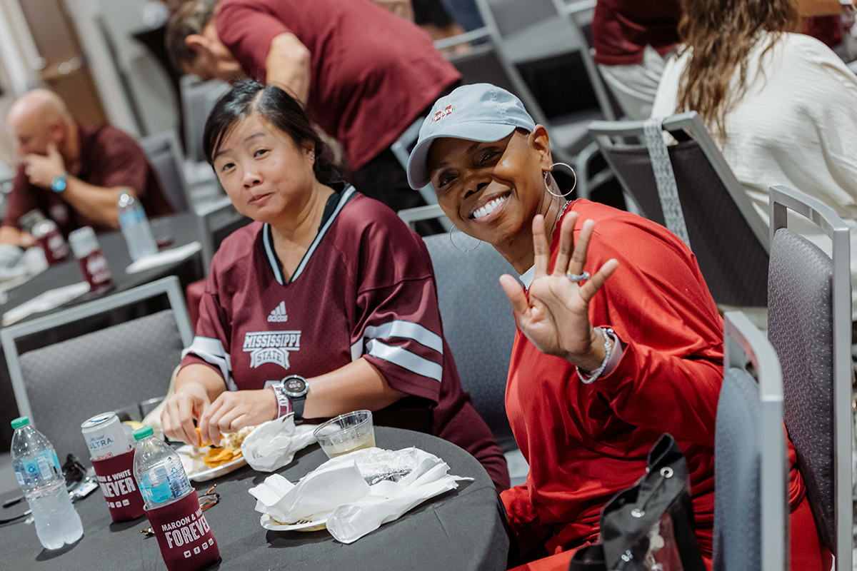 Two people sit at a table with food and drinks. The person on the right waves and smiles, wearing a red shirt and cap. The person on the left looks towards the camera, wearing a maroon jersey.