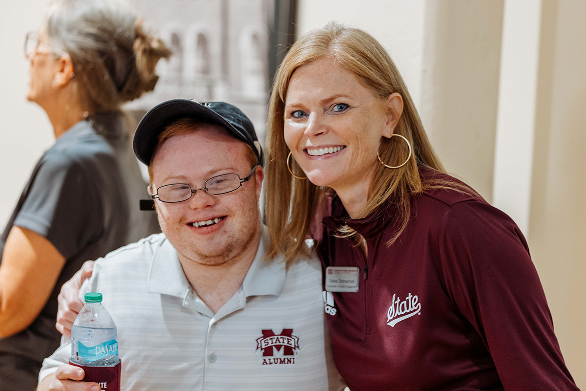 A woman in a maroon shirt poses with a smiling man wearing a cap and holding a water bottle. Both are in a bright indoor space with a blurred background.