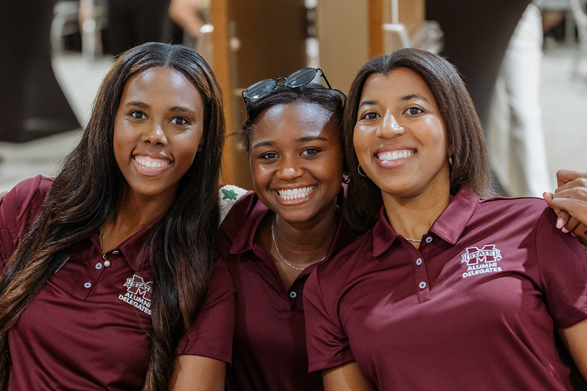 Three people smiling, wearing maroon polo shirts with a logo, standing together indoors in a casual group pose.