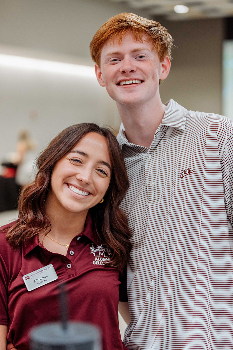 Two people smiling, one wearing a burgundy shirt with a name tag, and the other in a striped shirt, standing indoors.