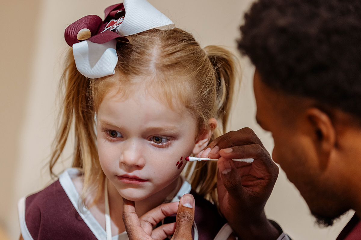 A young girl with pigtails and a maroon bow gets her face painted with a small maroon design by an adult using a swab.