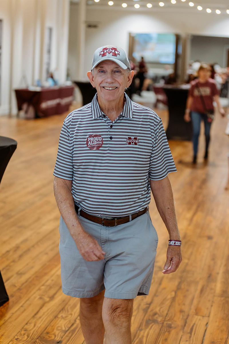 Elderly man wearing a cap and shorts walks in a large indoor hall with wooden flooring and other people in the background.