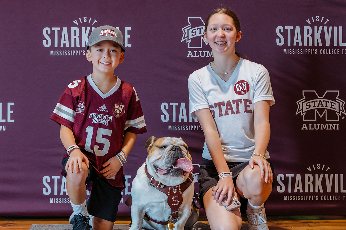 Two children in Mississippi State attire pose with a bulldog in front of a maroon backdrop that reads "Visit Starkville.