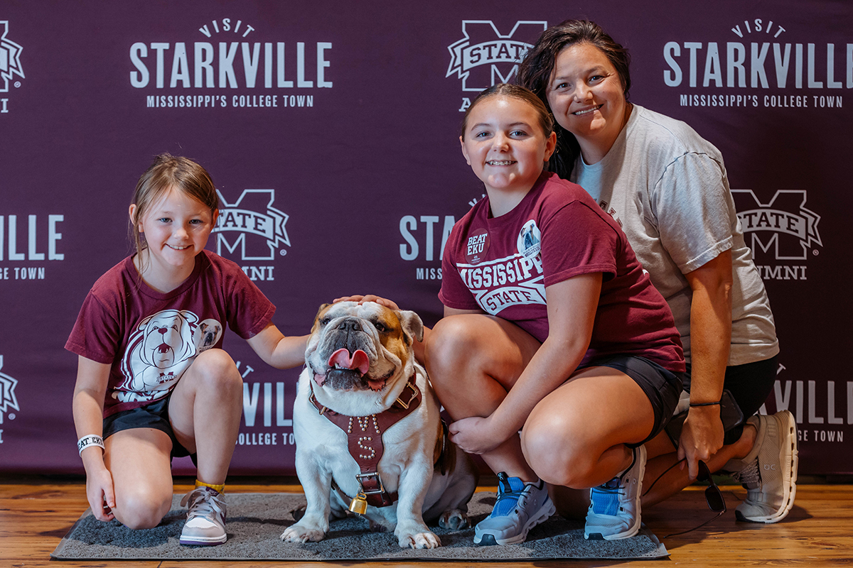 A bulldog sits with three people smiling beside it in front of a backdrop with "Visit Starkville" and "Mississippi State" logos.