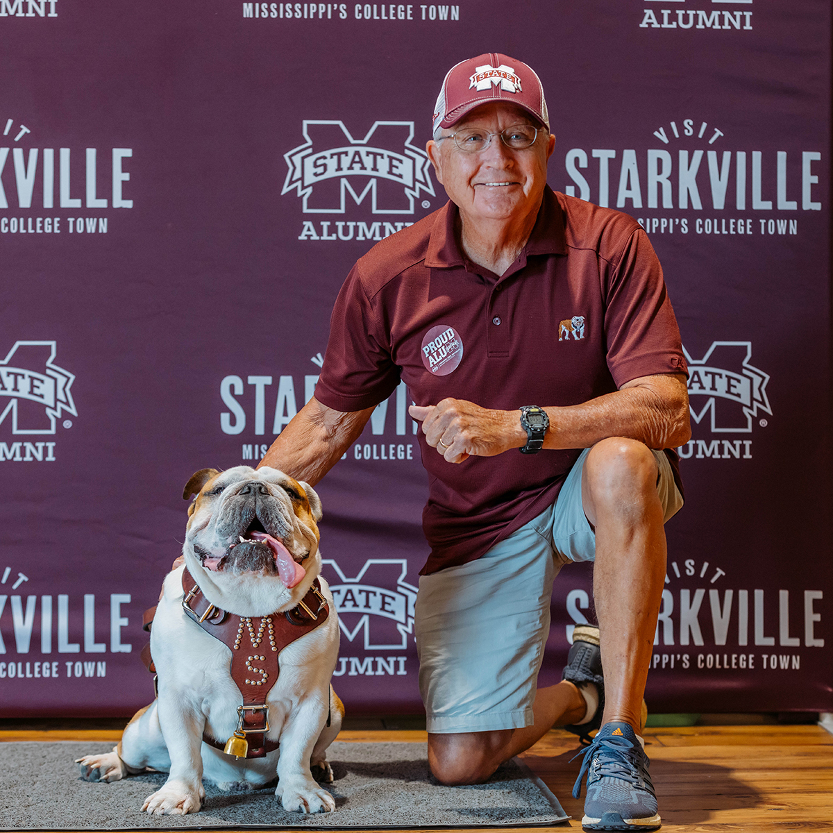 Man in maroon shirt and cap kneeling beside a bulldog wearing a harness. Background features a promotional backdrop with logos and "Mississippi’s College Town" text.
