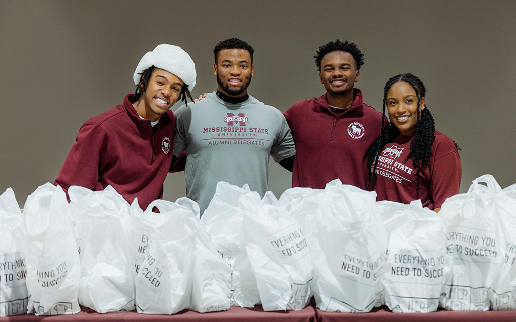 Four young adults stand behind a table filled with white bags labeled "Everything You Need To Succeed," wearing Mississippi State University apparel and smiling at the camera.