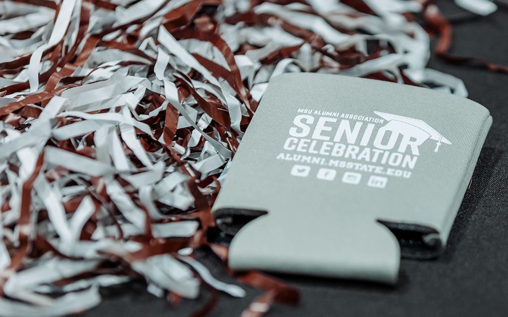 A foam drink holder with "Senior Celebration" printed on it sits next to maroon and white pom-poms on a black surface.