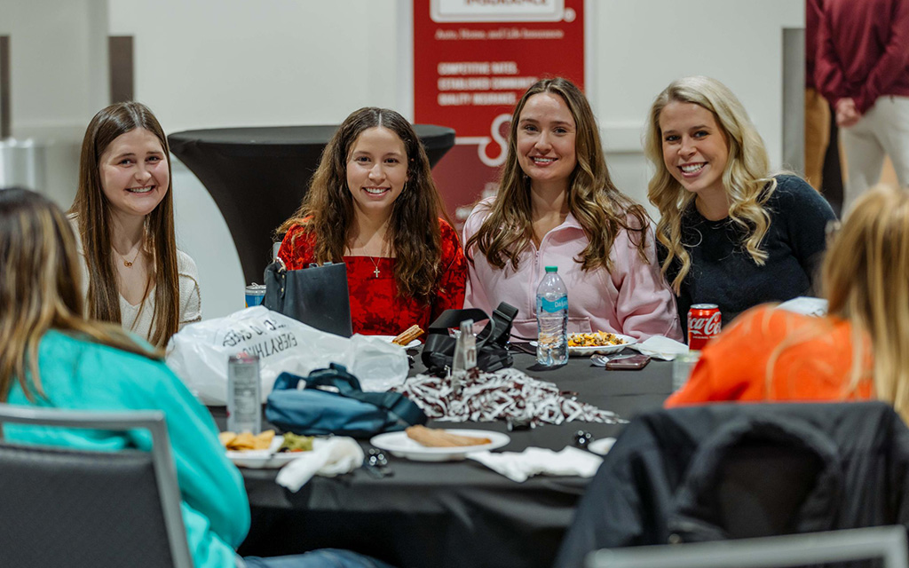 Four women are seated at a round table with food, drinks, and bags, smiling at the camera in an indoor event setting.
