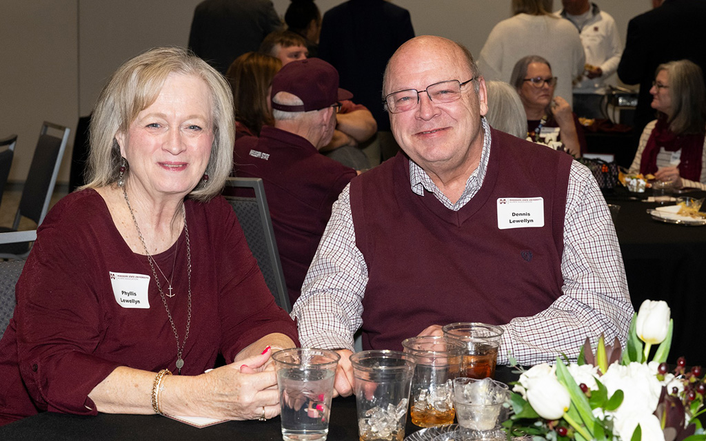 Two people sit at a table with drinks and flowers, wearing name tags that read "Phyllis" and "Dennis," at a social gathering with other attendees in the background.