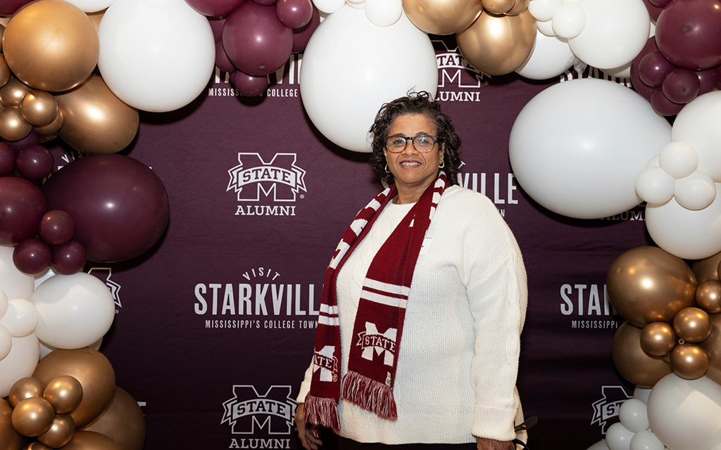 A woman wearing a white sweater and maroon scarf stands in front of a Mississippi State University Alumni backdrop with balloon decorations.