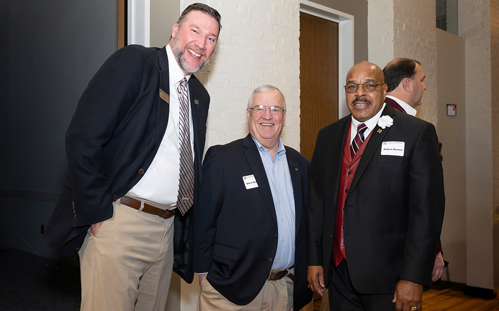 Three men wearing business attire stand indoors, smiling at the camera during an event. Each has a name tag on their jacket.