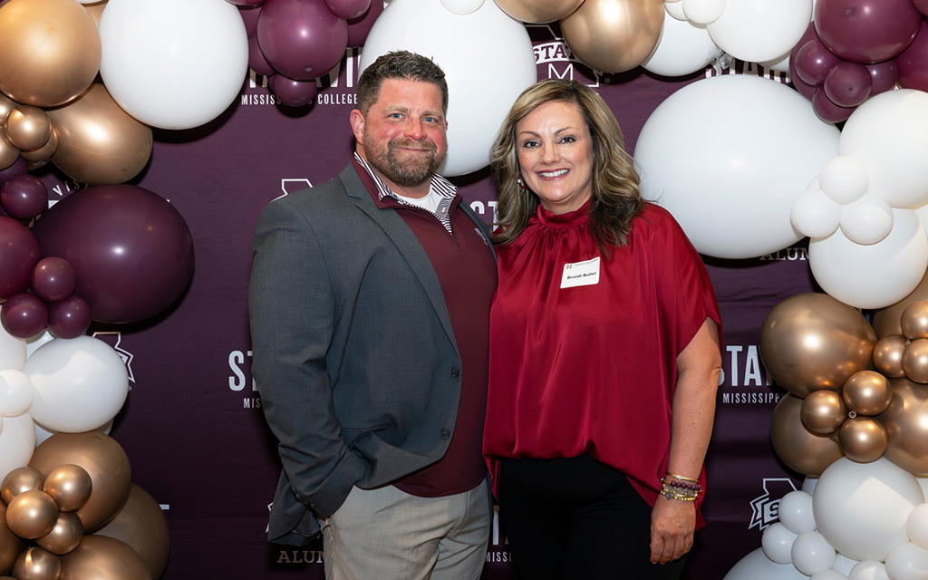 A man in a gray suit and a woman in a red blouse stand in front of a maroon photo backdrop with balloons at an event.