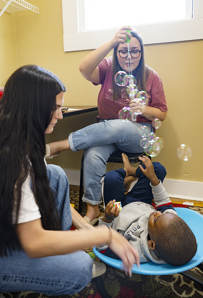 Two women interact with a young child who is lying in a blue bowl-shaped seat while reaching for bubbles being blown in a brightly lit room.