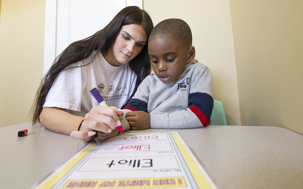 A woman helps a young boy write and solve addition equations on a worksheet at a table.