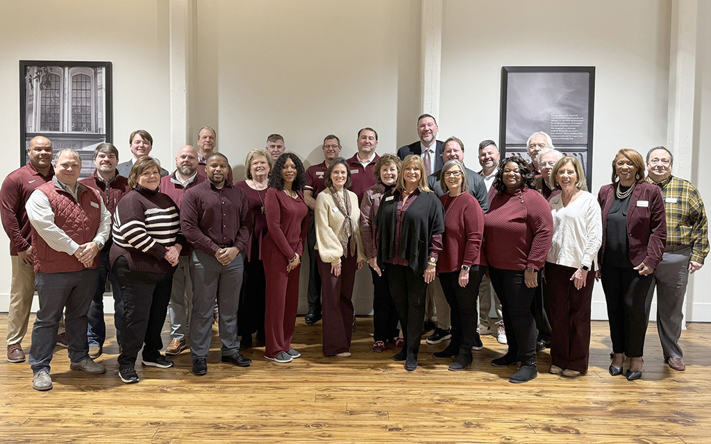 A group of about 27 adults pose together indoors in two rows, dressed in business casual attire, with neutral-colored walls and framed photos in the background.