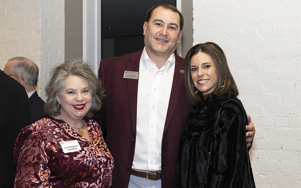 Three adults stand together at an indoor event, smiling at the camera. The man in the center wears a maroon blazer and the two women stand on either side of him.