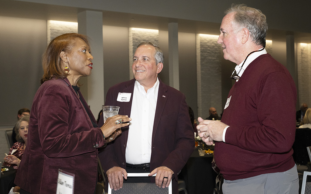 Three adults in business attire stand and converse at an indoor event; two men wear maroon sweaters and a woman holds a drink.