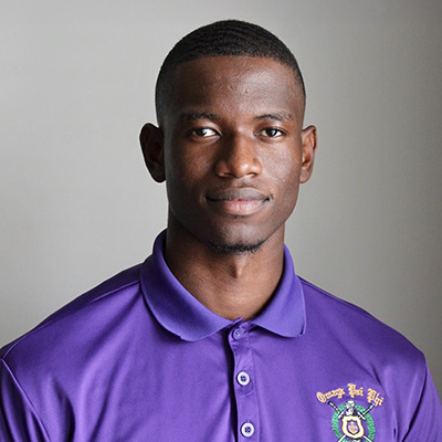 A man wearing a purple collared shirt with a crest logo stands against a plain gray background, looking at the camera with a neutral expression.
