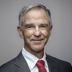 A middle-aged man in a gray suit, white shirt, and red patterned tie poses against a plain gray background, smiling slightly at the camera.