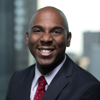 A man in a dark suit and red polka dot tie smiles at the camera, with a blurred cityscape visible in the background.