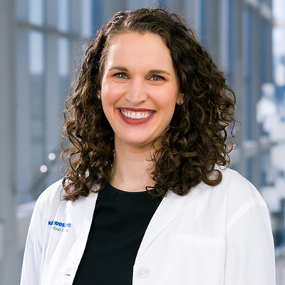 A woman with curly brown hair wearing a white lab coat and black top, smiling, stands in a bright hallway with large windows.