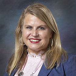 A woman with long blonde hair wearing a blue blazer, pink lace top, and hoop earrings, smiling in front of a dark, blurred background.