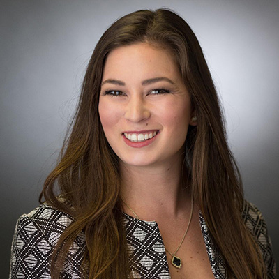 A woman with long brown hair wearing a black-and-white patterned jacket and a necklace, smiling in front of a gray gradient background.