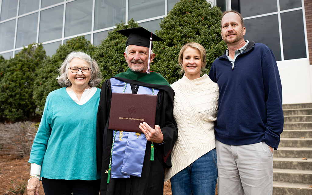 Four people stand together outside; the man in the center wears graduation robes and holds a diploma, flanked by three smiling adults.