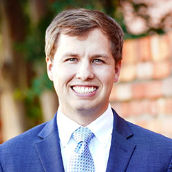 A man in a blue suit and patterned tie smiles at the camera with a brick wall and greenery in the background.