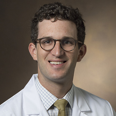 A man with short curly hair, glasses, and a white medical coat smiles at the camera against a neutral background.