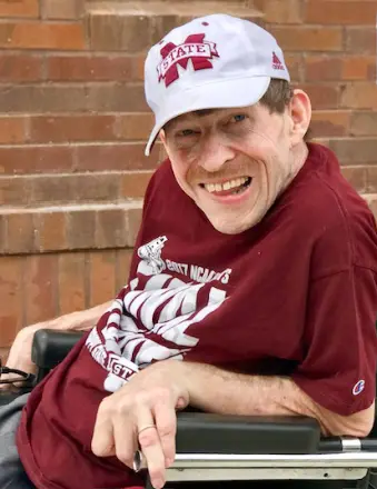 A man in a wheelchair wearing a maroon Mississippi State shirt and a matching cap smiles while sitting in front of brick steps.