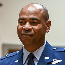A man in a blue military uniform with insignia is standing indoors, facing the camera, and smiling slightly.