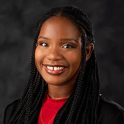 A woman with long braided hair, wearing a red top and black blazer, smiles at the camera against a dark, blurred background.