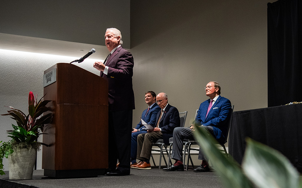 A man speaks at a podium while four men sit on chairs to his right on a stage in a conference room.