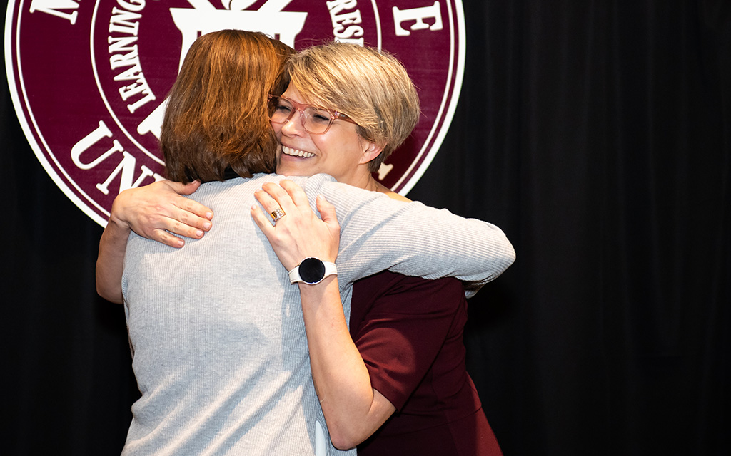 Two people hug in front of a maroon and white university seal, with one person smiling and wearing glasses and a smartwatch.