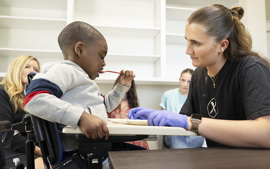 A young child in a wheelchair uses a straw while a woman in gloves assists him at a table. Three other women are seated in the background.