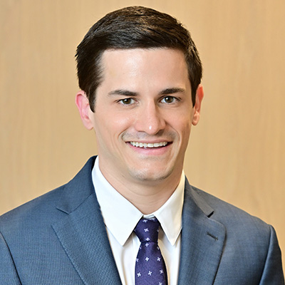 A man with short dark hair, wearing a blue suit, white shirt, and patterned tie, smiling in front of a plain light-colored background.