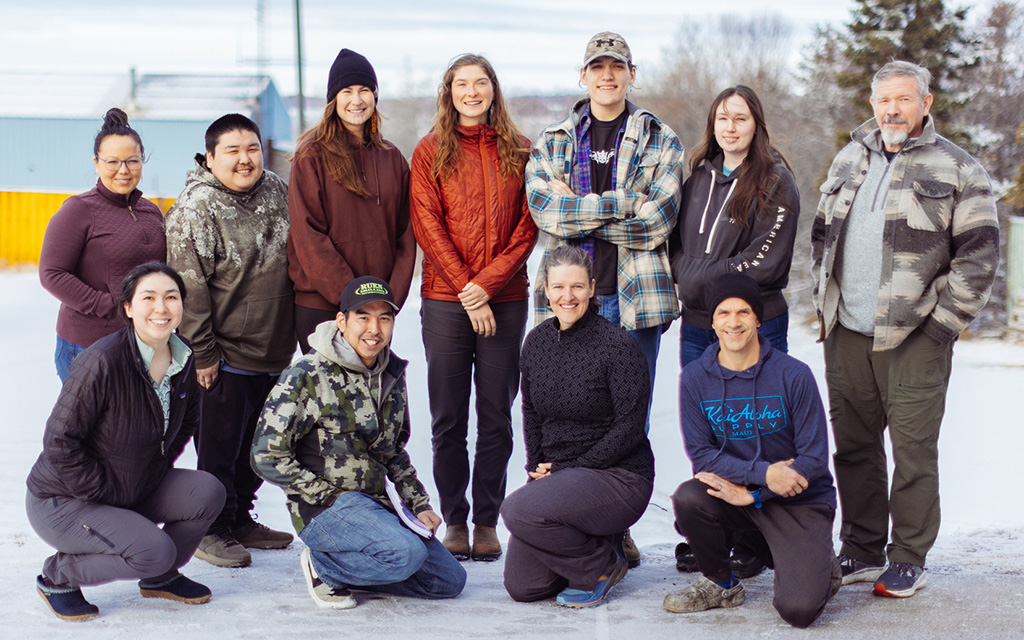 Eleven people pose outdoors on a cloudy day, some standing and some kneeling, dressed in casual, warm clothing. Trees and a building are visible in the background.