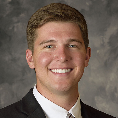 A young man in a dark suit, white shirt, and striped tie smiles at the camera in front of a mottled gray background.
